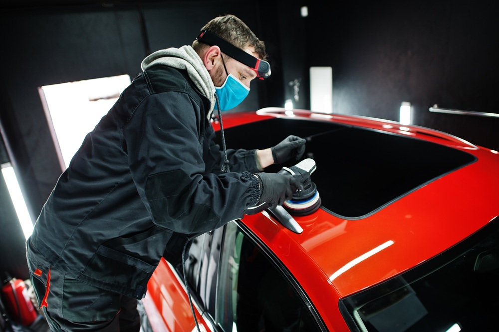 A man wearing a mask is polishing a car, focusing on the vehicle's surface for a shiny finish.